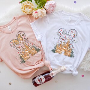 K&ouml;nnte beinhalten: Zwei T-Shirts, eines in blassem Rosa und eines in Wei&szlig;, jeweils mit einem skurrilen Design. Das Design umfasst ein Schloss, Feuerwerk und eine Mickey-Mouse-Kopfform. Eine Flasche Sekt befindet sich im Vordergrund.