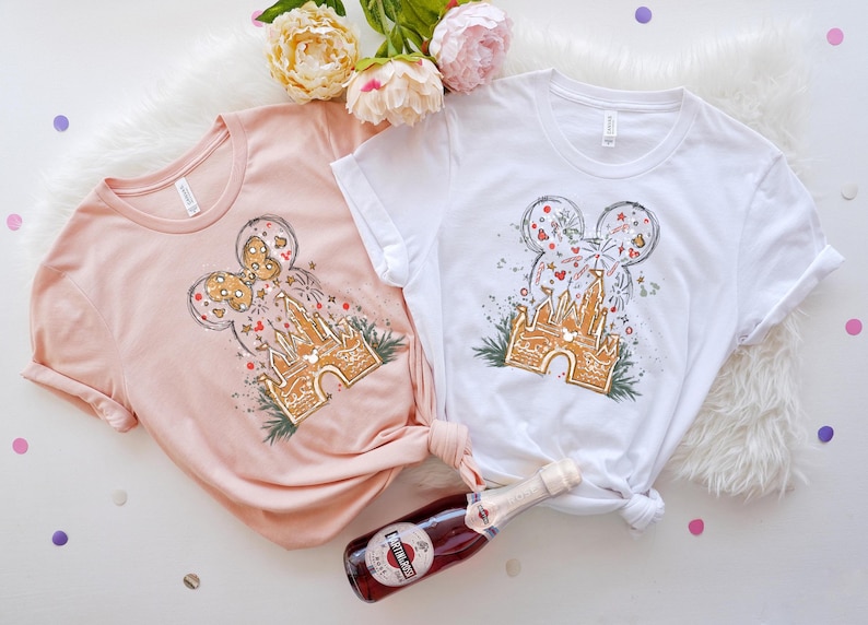 K&ouml;nnte beinhalten: Zwei T-Shirts, eines in blassem Rosa und eines in Wei&szlig;, jeweils mit einem skurrilen Design. Das Design umfasst ein Schloss, Feuerwerk und eine Mickey-Mouse-Kopfform. Eine Flasche Sekt befindet sich im Vordergrund.