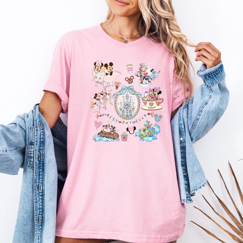 May include: Pink t-shirt featuring a colorful graphic with various Disney characters and the text "Best Day Ever" in a decorative frame. The design includes Mickey and Minnie Mouse, a castle, and other themed elements.