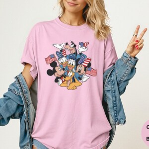 May include: Pink t-shirt featuring a graphic of Mickey Mouse, Minnie Mouse, Donald Duck, Goofy, and Pluto with American flags. The shirt is a Comfort Colors Blossom. The person is wearing a denim jacket.