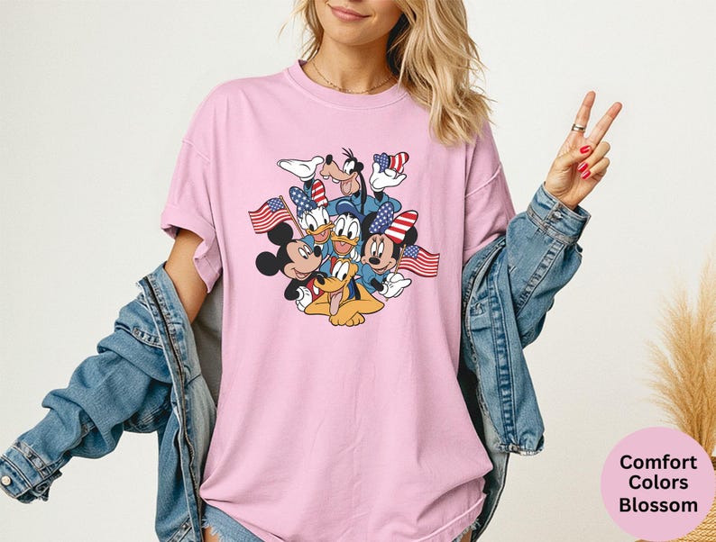 May include: Pink t-shirt featuring a graphic of Mickey Mouse, Minnie Mouse, Donald Duck, Goofy, and Pluto with American flags. The shirt is a Comfort Colors Blossom. The person is wearing a denim jacket.