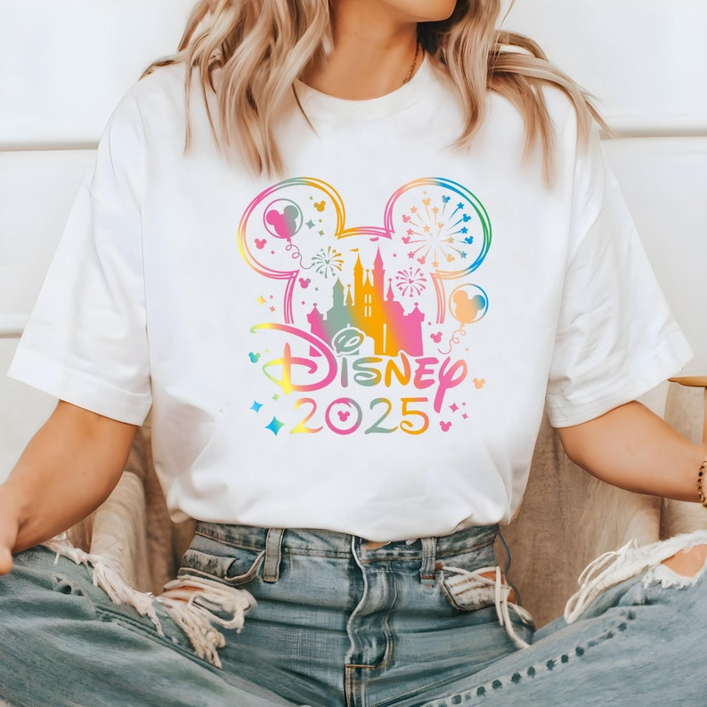 May include: White t-shirt with a rainbow-colored Mickey Mouse head design featuring a silhouette of Cinderella's castle and fireworks. The text "DISNEY 2025" is printed below the Mickey Mouse head.