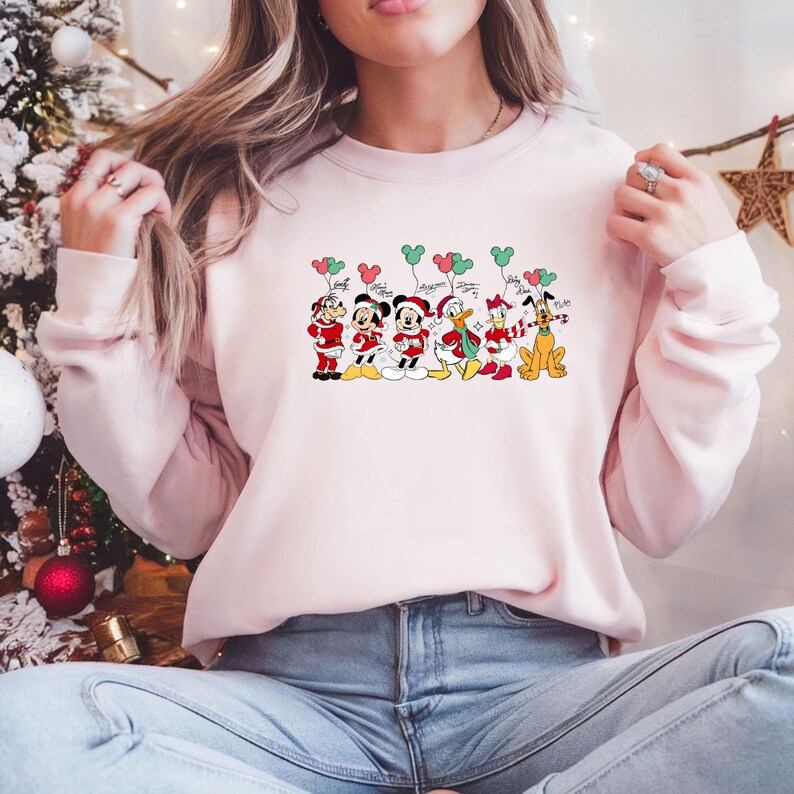 May include: A light pink sweatshirt featuring a festive graphic of Disney characters in Christmas attire. Mickey, Minnie, Donald, Daisy, Pluto, and Goofy are depicted with balloons. The design includes red, white, green, and black colors.