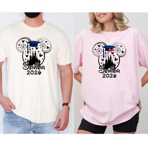 Puede incluir: Camisetas crema y rosa con un dise&ntilde;o negro de la cabeza de Mickey Mouse, un birrete, un castillo y fuegos artificiales. El texto "Senior 2026" est&aacute; debajo. Las camisetas las llevan dos personas.