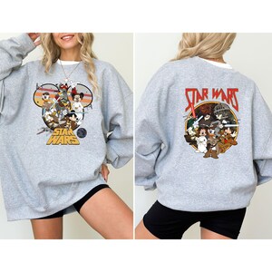 May include: A light gray sweatshirt featuring a Star Wars graphic with Disney characters on the front and back. The front graphic is in the shape of Mickey Mouse ears. The back graphic has the words "Star Wars" in red.