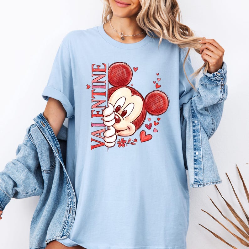 May include: Pale blue t-shirt with a Valentine's Day graphic. The design features a cartoon mouse with red ears and a red and white face, alongside the word "VALENTINE" in red vertical text. Small red hearts and flowers are also included.