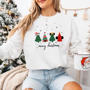 May include: White sweatshirt featuring four Christmas trees with Mickey Mouse ears and a Santa hat. The trees are decorated with red, green, and black accents. The words "merry christmas" are written in a script font below the trees.