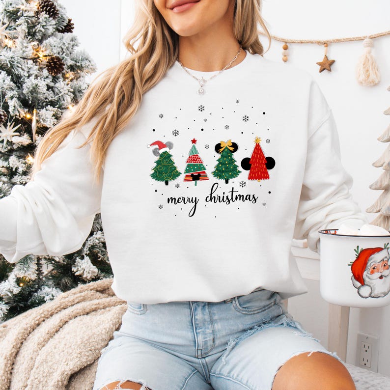 May include: White sweatshirt featuring four Christmas trees with Mickey Mouse ears and a Santa hat. The trees are decorated with red, green, and black accents. The words "merry christmas" are written in a script font below the trees.