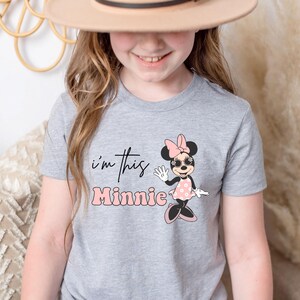 K&ouml;nnte beinhalten: Ein graues T-Shirt mit einem Minnie Mouse-Motiv. Das Design zeigt Minnie in einem rosa gepunkteten Kleid, Sonnenbrille und Schleife. Der Text "i'm this Minnie" ist oberhalb und unterhalb des Minnie Mouse-Bildes aufgedruckt.