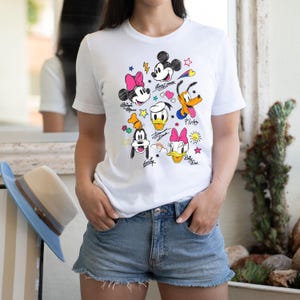 May include: White t-shirt with a colorful cartoon design featuring Mickey Mouse, Minnie Mouse, Donald Duck, Goofy, and Pluto. The characters are drawn in a doodle style with colorful accents and their names written underneath.