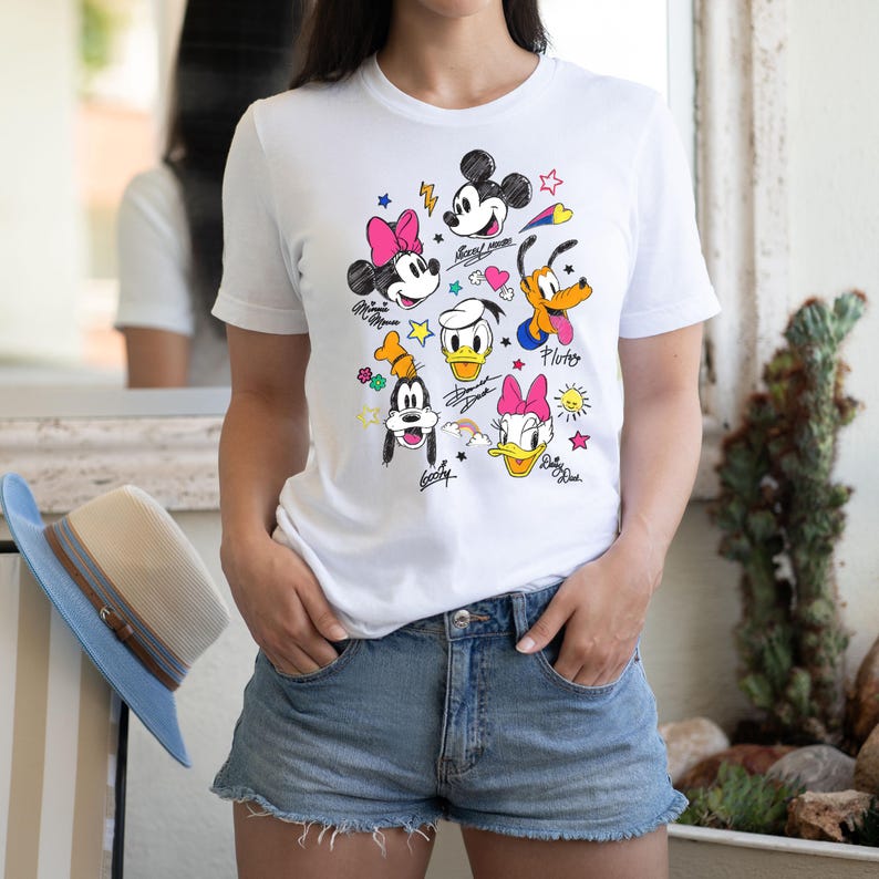 May include: White t-shirt with a colorful cartoon design featuring Mickey Mouse, Minnie Mouse, Donald Duck, Goofy, and Pluto. The characters are drawn in a doodle style with colorful accents and their names written underneath.