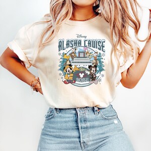 Pu&ograve; includere: T-shirt color crema con un design Disney Alaska Cruise. La grafica include Topolino, Minnie, Paperino e Pippo su una nave da crociera, con la scritta "Alaska Cruise 2025". Il design &egrave; circondato da fiocchi di neve e pini.