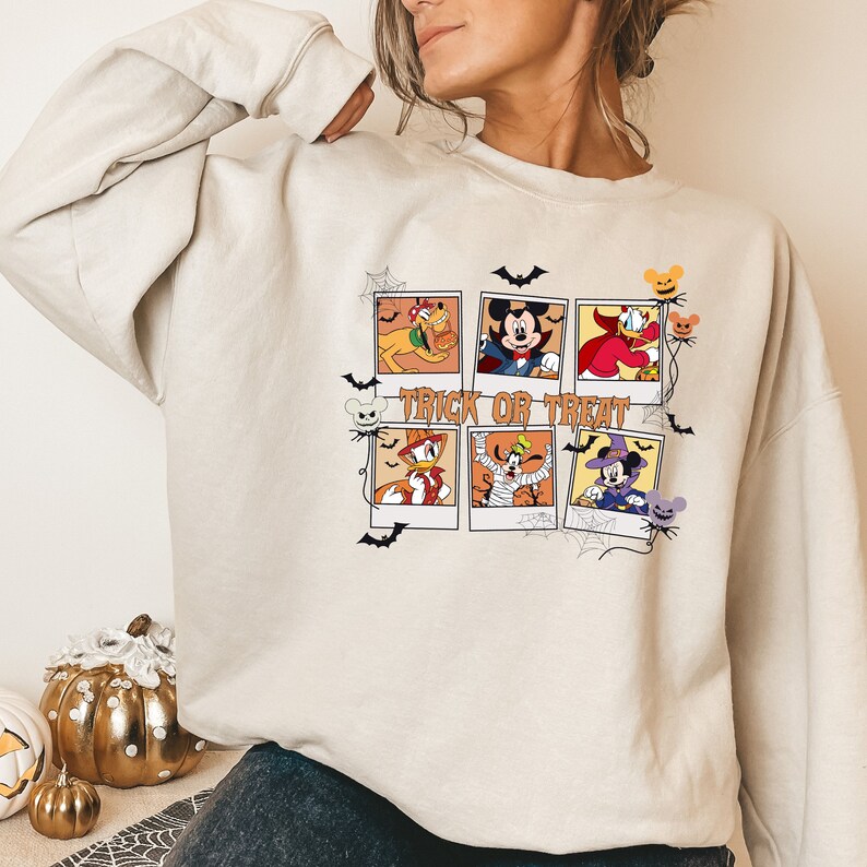 Op de afbeelding: Een cr&egrave;mekleurige trui met een grafische print van Mickey Mouse en andere Disney-figuren die verkleed zijn in Halloween-kostuums. De afbeelding bevat de tekst "Trick or Treat".