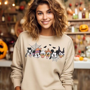 Puede incluir: Una sudadera de color beige con un gr&aacute;fico de Mickey Mouse, Minnie Mouse, Donald Duck, Goofy, Pluto y Mal&eacute;fica en un tema de Halloween. Los personajes est&aacute;n rodeados por una telara&ntilde;a y murci&eacute;lagos.