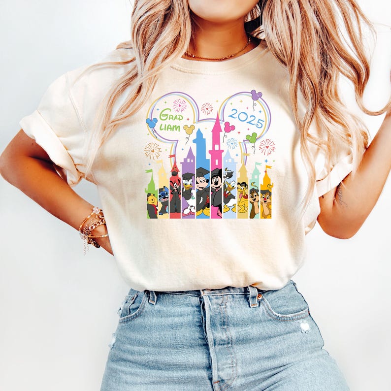 Puede incluir: Una camiseta blanca con un gr&aacute;fico colorido de Mickey Mouse y otros personajes de Disney frente a un castillo. El gr&aacute;fico incluye el texto "Grad Liam" y "2025".