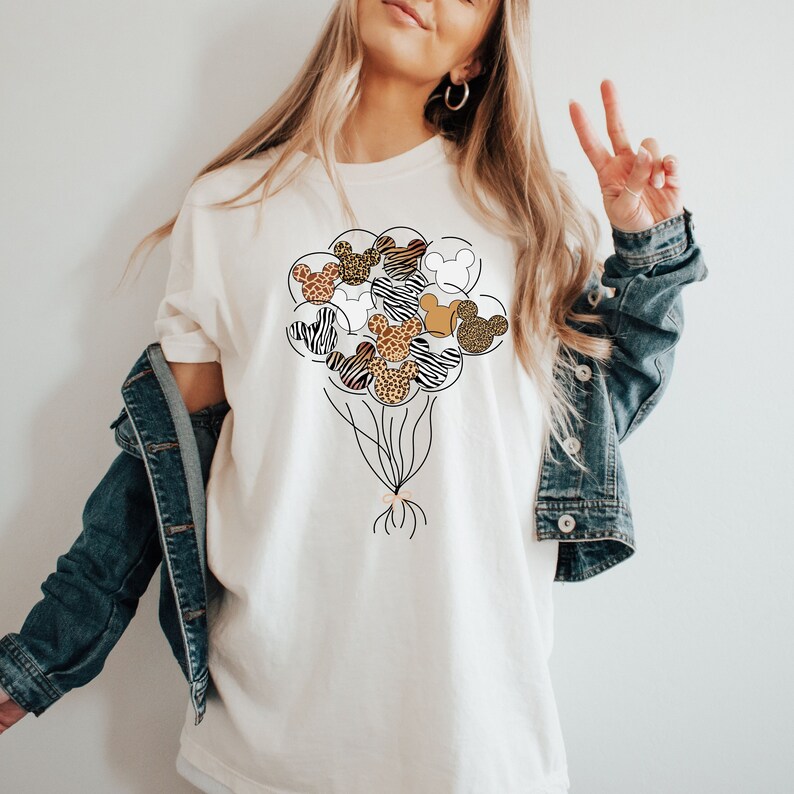 Puede incluir: Camiseta blanca con un gr&aacute;fico de un mont&oacute;n de cabezas de Mickey Mouse con estampado de animales en forma de globos.
