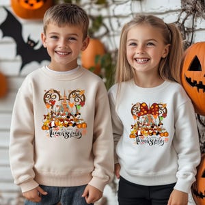 May include: Two children wearing cream and white sweatshirts with a Thanksgiving design. The design features Mickey Mouse and friends, a castle, pumpkins, and the words "Happy Thanksgiving". The background includes Halloween decorations.