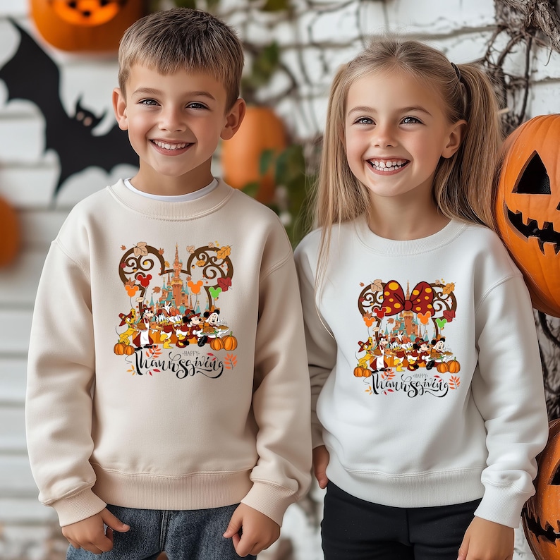May include: Two children wearing cream and white sweatshirts with a Thanksgiving design. The design features Mickey Mouse and friends, a castle, pumpkins, and the words "Happy Thanksgiving". The background includes Halloween decorations.