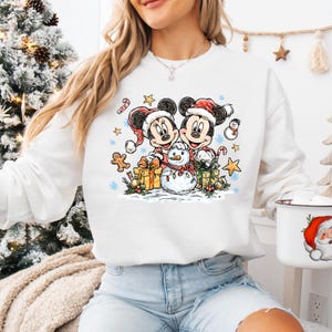 May include: White sweatshirt featuring a festive graphic of Mickey and Minnie Mouse in Santa hats, with a snowman, gifts, and candy canes. The design includes snowflakes and stars, creating a holiday theme.