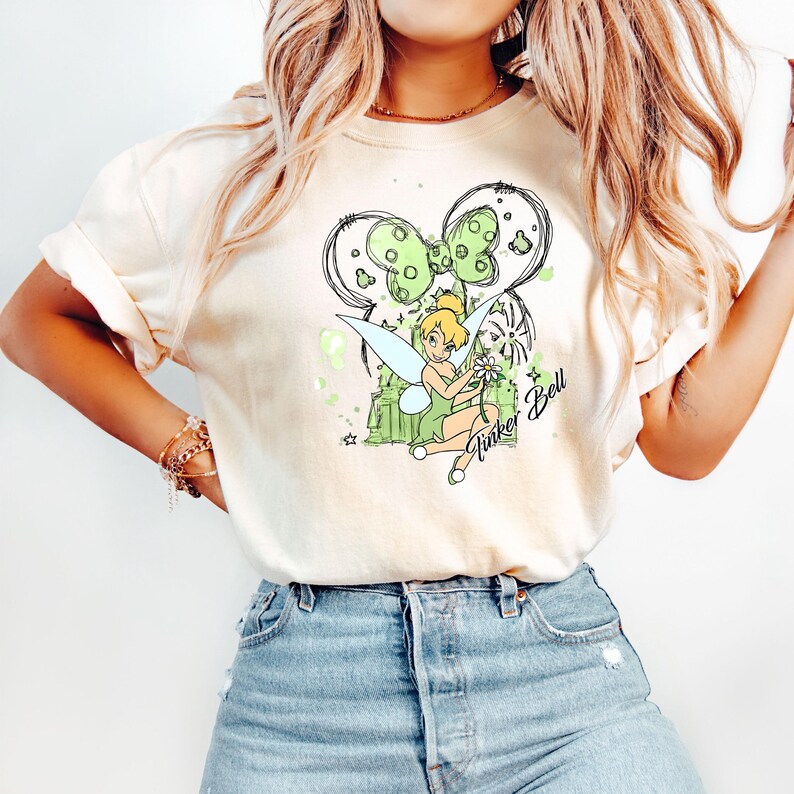 May include: A white t-shirt with a green and black illustration of Tinker Bell, a fairy from the Disney movie Peter Pan. The illustration includes a green bow, a green castle, and the text "Tinker Bell".