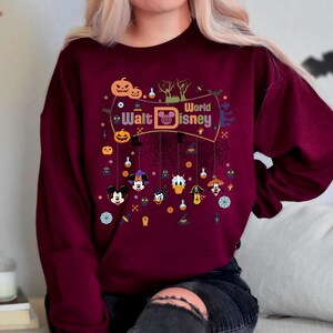 May include: A maroon sweatshirt with a Halloween-themed design featuring Mickey Mouse, Donald Duck, Goofy, and other Disney characters. The design includes the text "Walt Disney World" and various Halloween icons like pumpkins, bats, and ghosts.