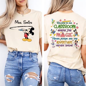 May include: A beige t-shirt with a Mickey Mouse illustration on the front and text that says "Mrs. Smith" above the illustration. The back of the shirt has a colorful design with Disney characters and the text "Welcome to our Classroom where the magic never stops and the adventure never ends!"