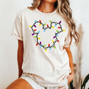 May include: A cream colored t-shirt with a design of colorful Christmas lights in the shape of Mickey Mouse ears.