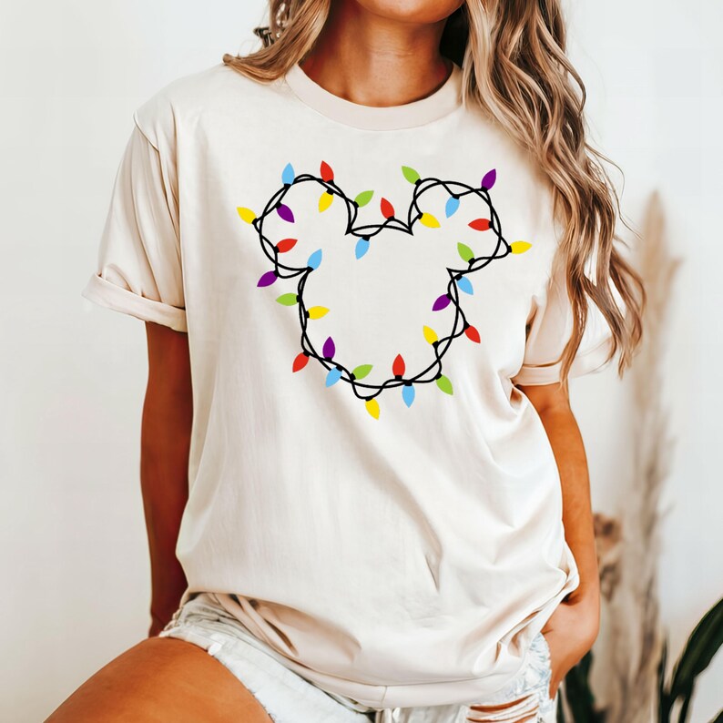 May include: A cream colored t-shirt with a design of colorful Christmas lights in the shape of Mickey Mouse ears.