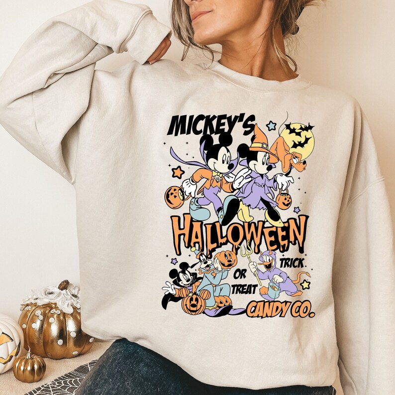 May include: A light beige sweatshirt with a graphic print of Mickey Mouse and Minnie Mouse dressed in Halloween costumes. The text "Mickey's Halloween Trick or Treat Candy Co." is printed on the sweatshirt.