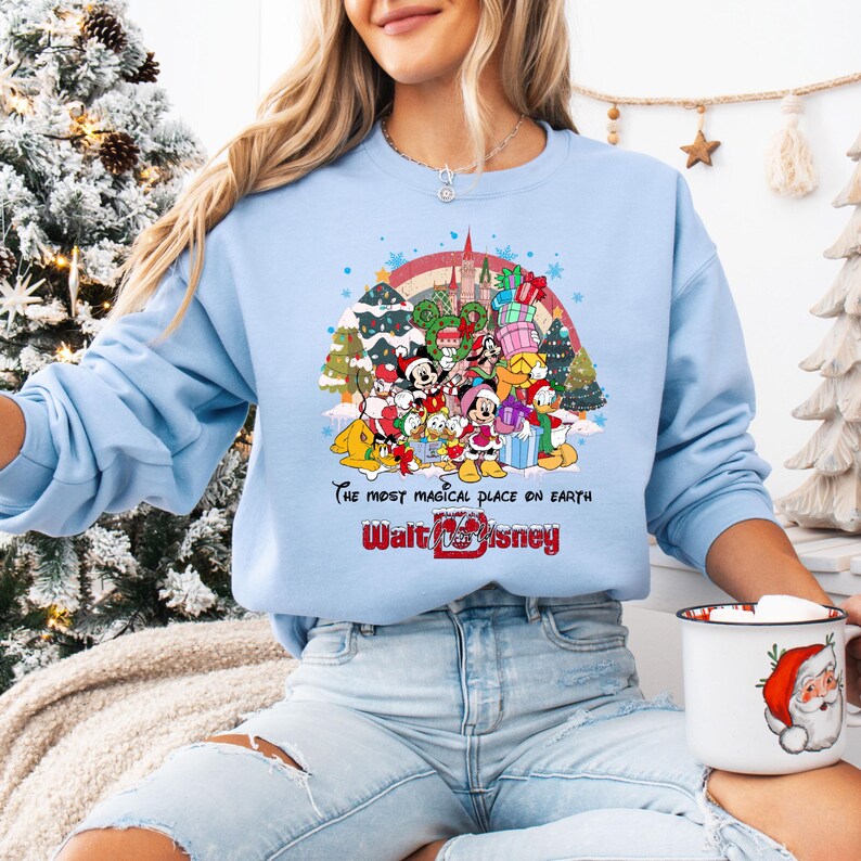 Op de afbeelding: Lichtblauw sweatshirt met een kleurrijke afbeelding van Disney-personages, een regenboog en een kasteel. De tekst "The most magical place on Earth" en "Walt Disney" staan eronder. Het sweatshirt heeft een ronde hals.