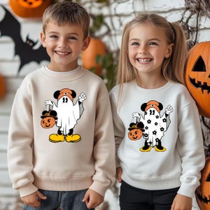 May include: Two children wearing Halloween-themed sweatshirts. The sweatshirts feature a cartoon ghost design with Mickey Mouse ears, holding a pumpkin-shaped treat bag. The background includes pumpkins and a bat silhouette.