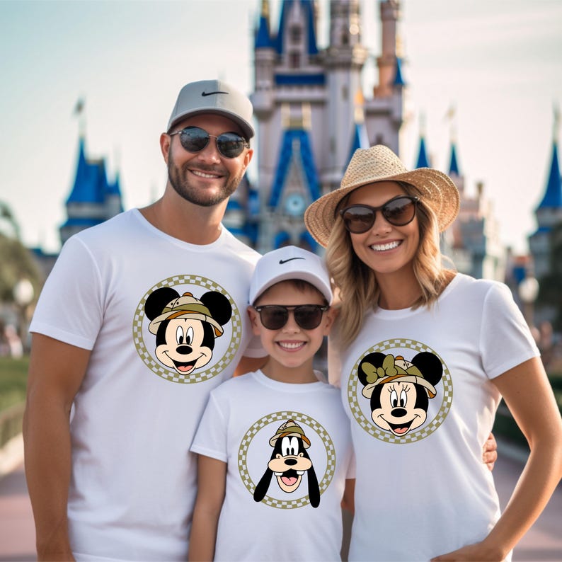 May include: A family of three wearing white t-shirts with cartoon character images. The father's shirt features Mickey Mouse wearing a safari hat, the mother's shirt features Minnie Mouse wearing a safari hat, and the child's shirt features Goofy wearing a safari hat.