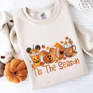 K&ouml;nnte beinhalten: Cremefarbenes Sweatshirt mit einem herbstlichen Motiv. Das Design umfasst einen Pumpkin Spice Latte, K&uuml;rbislaternen, einen Football und eine Tasse hei&szlig;e Schokolade. Der Text "Tis The Season" ist ebenfalls enthalten.