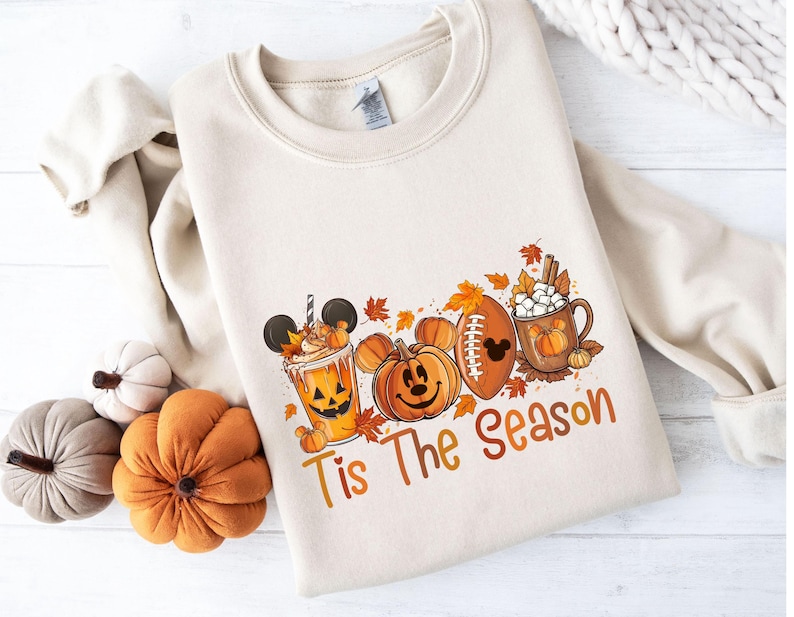 K&ouml;nnte beinhalten: Cremefarbenes Sweatshirt mit einem herbstlichen Motiv. Das Design umfasst einen Pumpkin Spice Latte, K&uuml;rbislaternen, einen Football und eine Tasse hei&szlig;e Schokolade. Der Text "Tis The Season" ist ebenfalls enthalten.