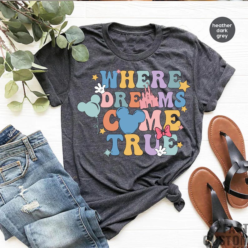 May include: A heather dark grey t-shirt with the text "Where Dreams Come True" in colorful letters. The text is surrounded by stars, Mickey Mouse ears, a castle, and a balloon.
