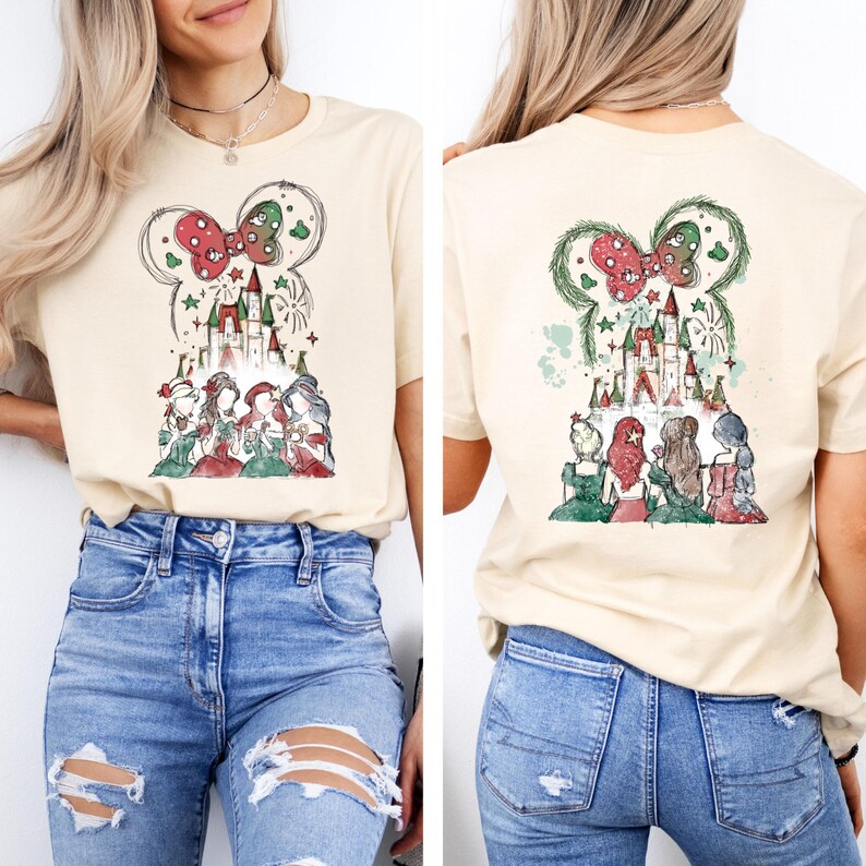May include: A beige t-shirt featuring a whimsical illustration of a castle, princesses, and a Mickey Mouse-shaped bow with Christmas accents. The design is repeated on the back. The shirt is paired with blue jeans.