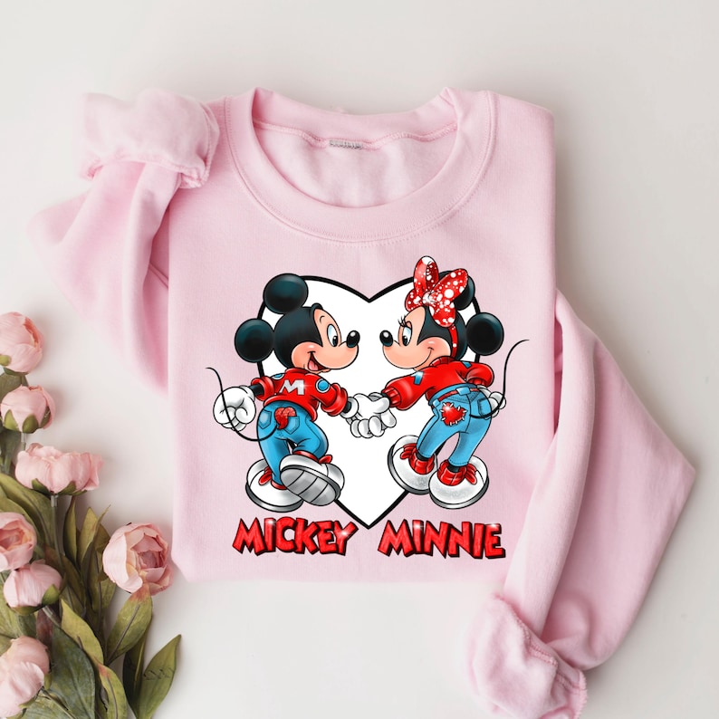 May include: Pink sweatshirt with a white heart design featuring Mickey Mouse and Minnie Mouse holding hands. The text "Mickey Minnie" is printed below the heart.