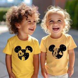 May include: Two children wearing yellow t-shirts with black Mickey Mouse designs. One shirt features Mickey giving a thumbs up, the other features Minnie Mouse. Both children are smiling.