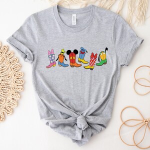 May include: A gray t-shirt featuring a colorful graphic of cartoon cowboy boots. The boots are designed with various patterns and character-inspired details, including a bow, Mickey Mouse ears, and polka dots. The shirt is knotted at the bottom.