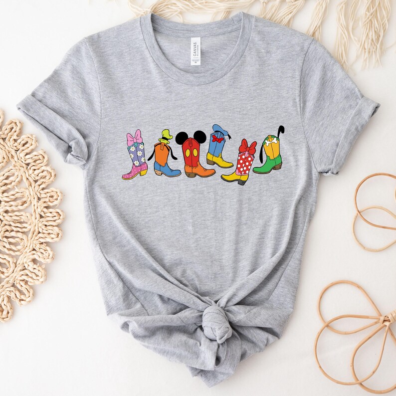 May include: A gray t-shirt featuring a colorful graphic of cartoon cowboy boots. The boots are designed with various patterns and character-inspired details, including a bow, Mickey Mouse ears, and polka dots. The shirt is knotted at the bottom.