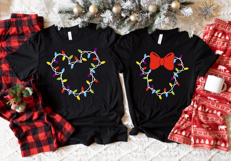 May include: Two black t-shirts with a Christmas lights design in the shape of Mickey Mouse and Minnie Mouse. The Mickey Mouse shirt has a white outline and the Minnie Mouse shirt has a red bow.