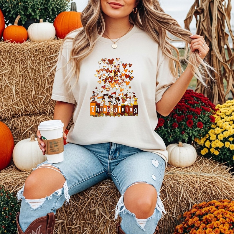 May include: A beige t-shirt with a Thanksgiving-themed graphic. The graphic features cartoon characters and heart-shaped balloons in shades of brown and orange. The word "Thanksgiving" is printed in a rectangular box. The person is wearing blue jeans and brown boots.