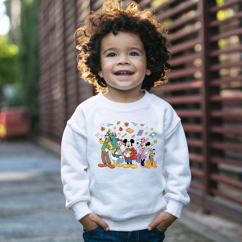 May include: A child wearing a white sweatshirt featuring a colorful graphic of Disney characters, including Mickey Mouse, Minnie Mouse, Donald Duck, and Goofy. The design includes autumn-themed elements like leaves and books. The child is smiling.
