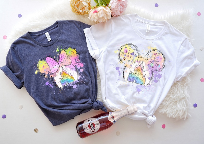 Puede incluir: Dos camisetas con un dise&ntilde;o de cabeza de Mickey Mouse de colores arco&iacute;ris. Una camiseta es azul marino y la otra es blanca. El dise&ntilde;o presenta un castillo y fuegos artificiales.