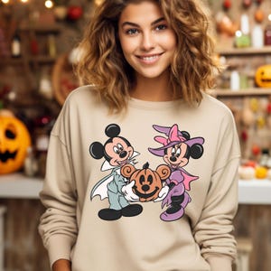 May include: A beige sweatshirt with a graphic print of Mickey Mouse and Minnie Mouse dressed in Halloween costumes. Mickey is dressed as a vampire and Minnie is dressed as a witch. They are holding a jack-o'-lantern.