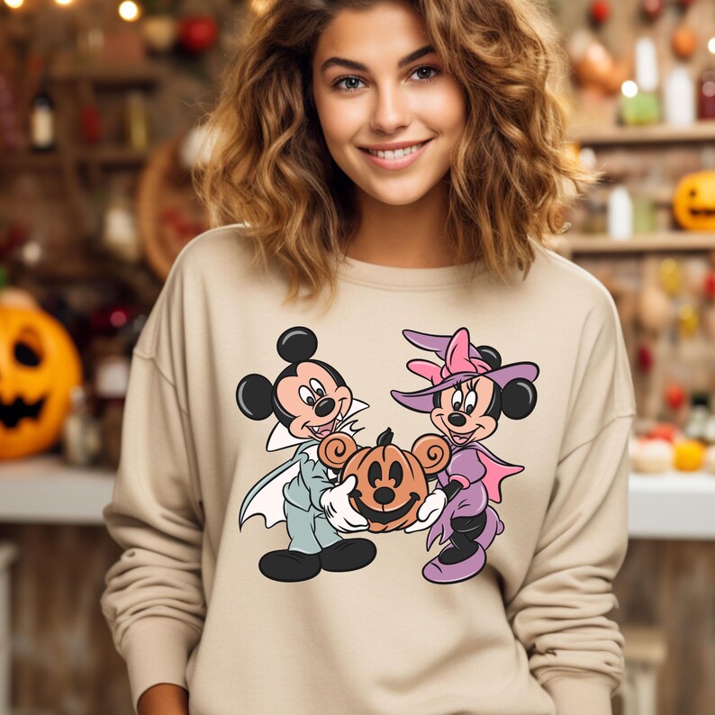May include: A beige sweatshirt with a graphic print of Mickey Mouse and Minnie Mouse dressed in Halloween costumes. Mickey is dressed as a vampire and Minnie is dressed as a witch. They are holding a jack-o'-lantern.