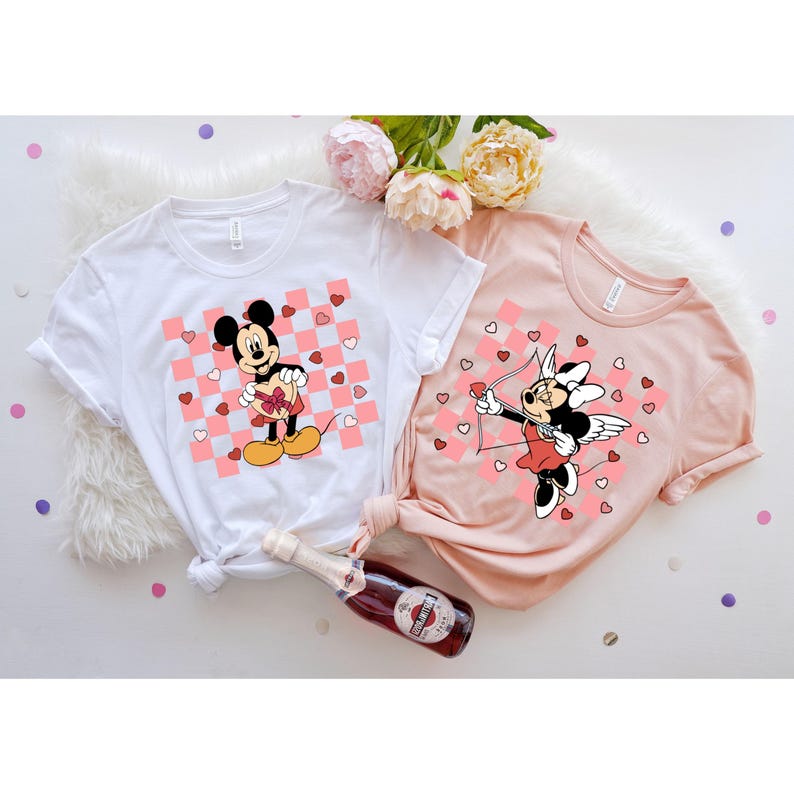 Pode incluir: Duas camisetas brancas com um padr&atilde;o xadrez rosa e branco. Uma camiseta apresenta o Mickey Mouse segurando uma caixa de chocolates em forma de cora&ccedil;&atilde;o. A outra camiseta apresenta a Minnie Mouse vestida como Cupido com asas e uma flecha.