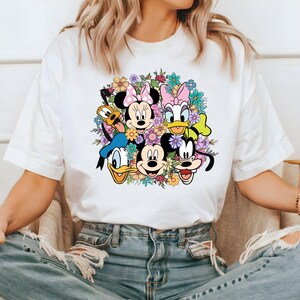 May include: White t-shirt with a colorful graphic print of Mickey Mouse, Minnie Mouse, Donald Duck, Goofy, and Pluto surrounded by flowers.