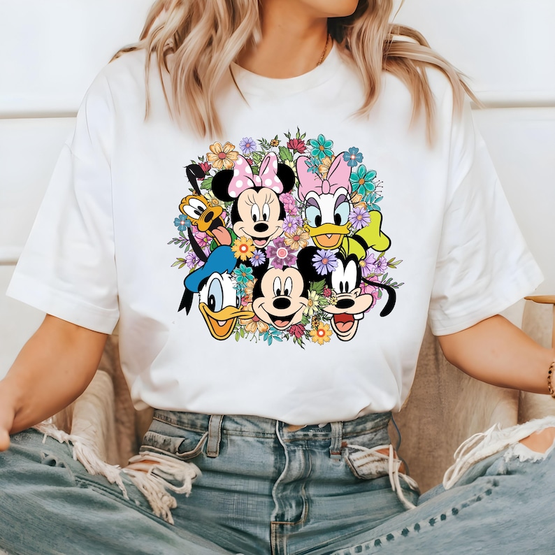 May include: White t-shirt with a colorful graphic print of Mickey Mouse, Minnie Mouse, Donald Duck, Goofy, and Pluto surrounded by flowers.