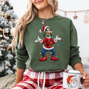 May include: An olive green sweatshirt with a cartoon Goofy character in a Santa hat and red Christmas attire, holding festive lights. The sweatshirt is worn with red and white plaid pyjama bottoms. A white mug with a Santa Claus design is also present.